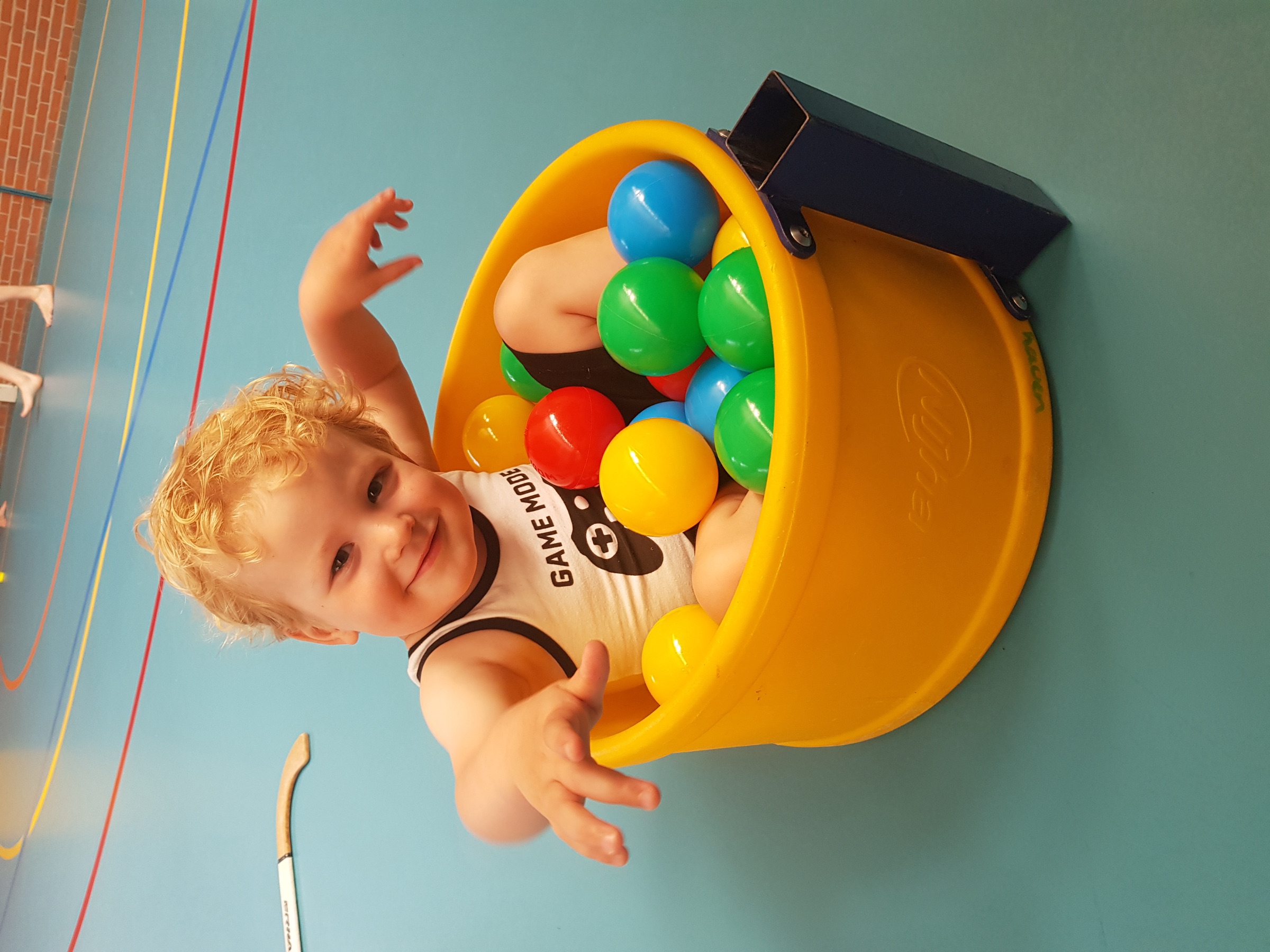 Kind in een gele ballenbak in de gymzaal, met ballen in de lucht