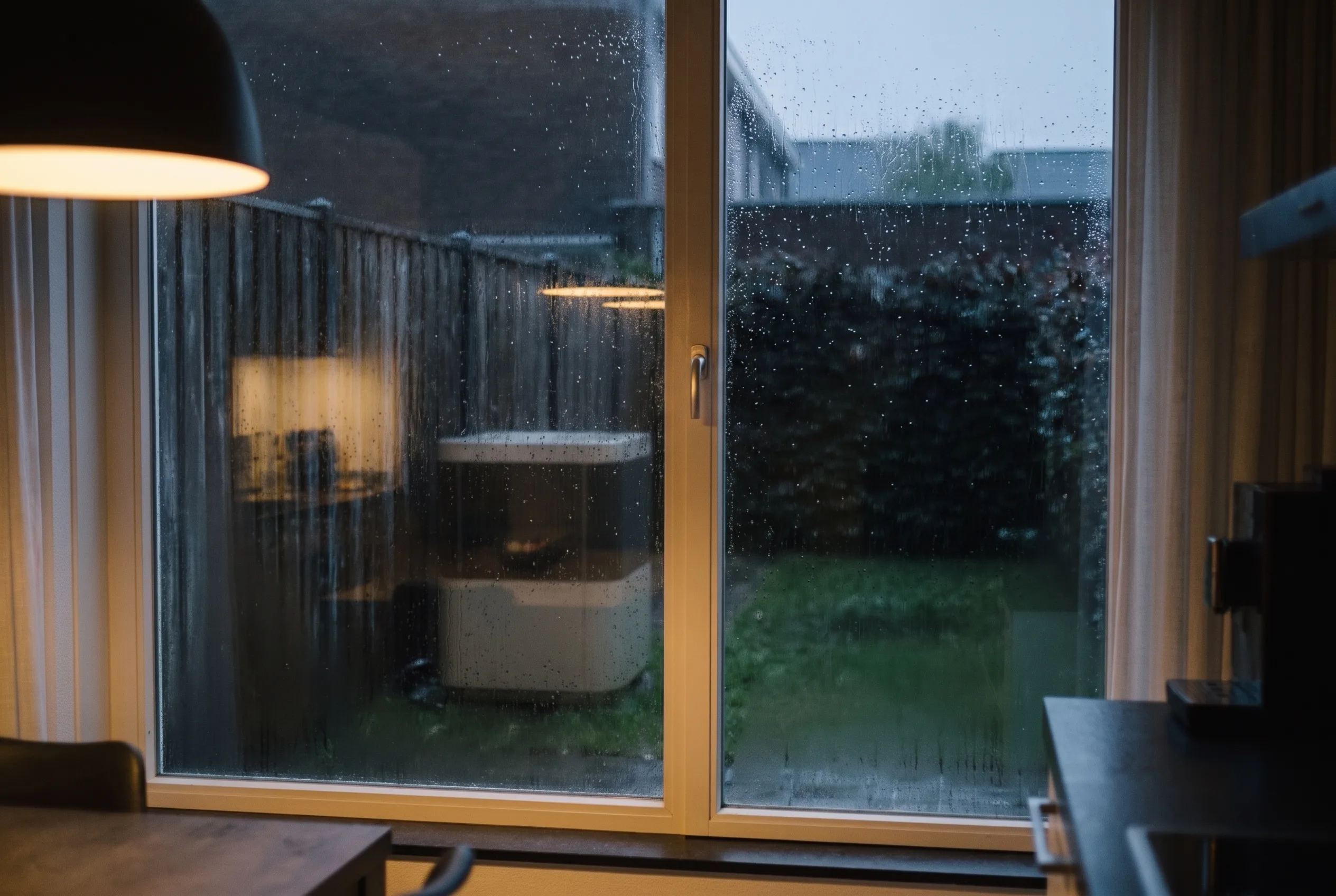Dezelfde tuin vanuit de keuken gezien, door een raam met regenstrepen, in het donker.